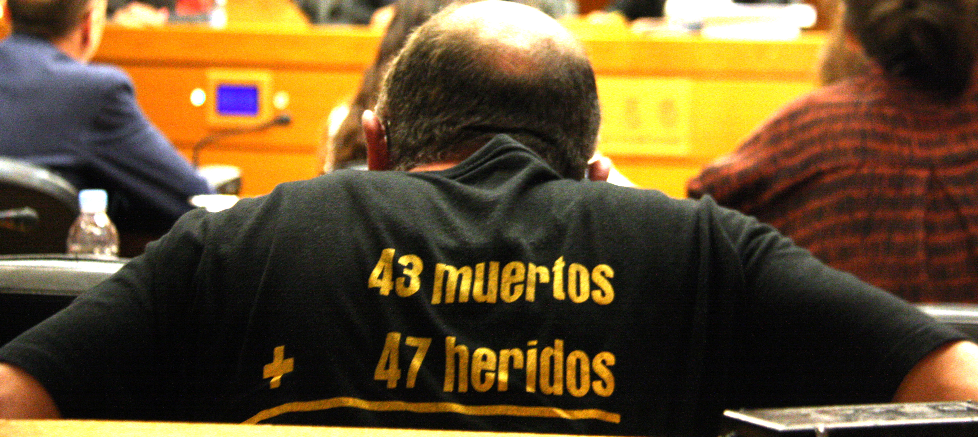 Un familiar de les víctimes del metro de València d'esquenes amb una samarreta que recorda el número de morts i ferits de l'accident. Al fons i desenfocat, Juan Cotino a la comissió d'investigació (fotografia d'ACN).