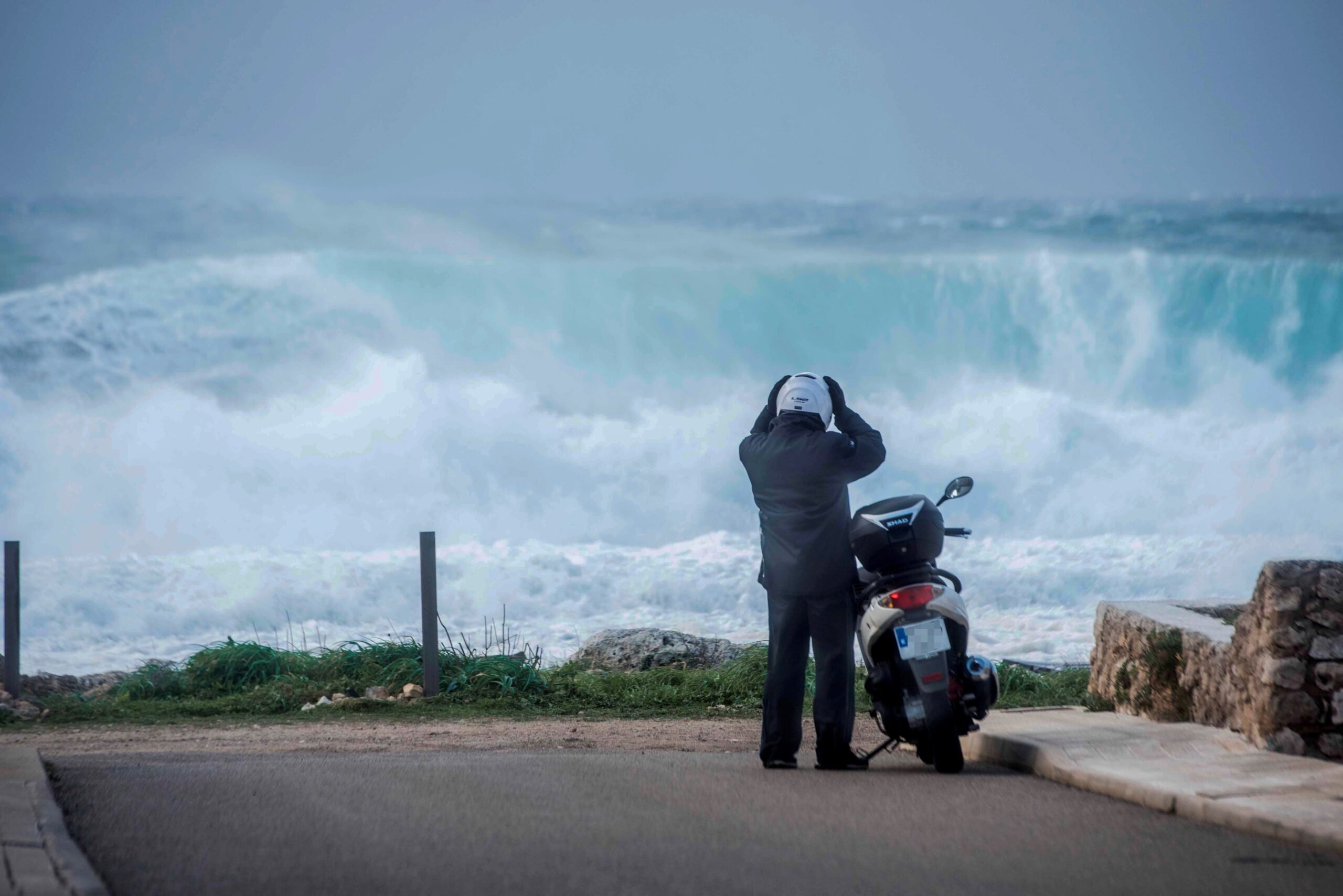 Un motorista es prepara per muntar en el seu scooter mentre grans onades trenquen a Biniancolla (Menorca). Fotografia: EFE/ David Arquimnau Sintes