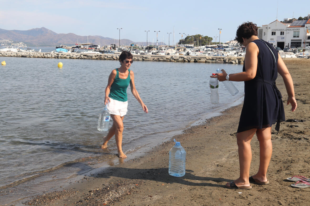 Dues dones omplen ampolles i garrafes del mar a la platja del Port de la Selva