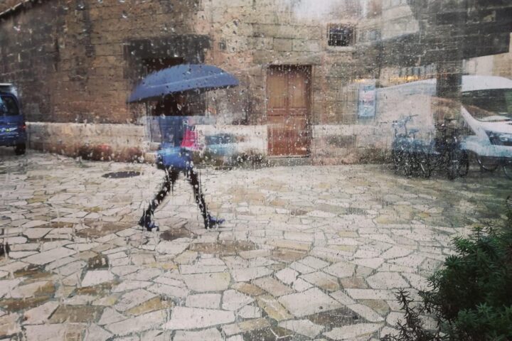 Una dona passeja pel centre de Palma amb paraigua, vista a través d’un vidre amb gotes de pluja.